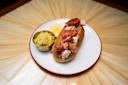 A lobster roll with chunks of lobster meat in a bun is served on a white plate with a lemon wedge and a small cup of coleslaw. The plate is on a wooden table with a sunburst pattern.