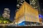 A large, cube-shaped building with a golden, illuminated exterior stands among tall skyscrapers at night, with streetlights and trees lining the sidewalk in front.