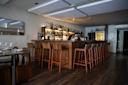 A bartender prepares drinks behind a wooden bar in a modern, dimly lit restaurant with high wooden stools, shelves of bottles, and set dining tables.