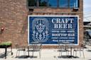 Outdoor seating area with metal tables and chairs in front of a brick wall featuring a large blue and white mural advertising craft beer, a rooftop bar, market, live music, games, and private event spaces.