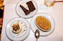 Three desserts on white plates: a slice of chocolate cake with whipped cream, a crème brûlée with a spoon, and a panna cotta topped with caramel sauce and nuts, next to a glass of water on a white table.