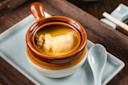 A close-up of a brown and white ceramic bowl with a handle, filled with soup containing a large piece of seafood, possibly abalone. The bowl is placed on a rectangular white plate with a ceramic spoon beside it.