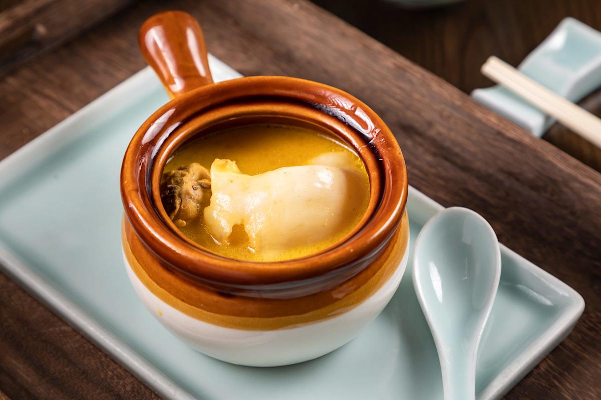 A close-up of a brown and white ceramic bowl with a handle, filled with soup containing a large piece of seafood, possibly abalone. The bowl is placed on a rectangular white plate with a ceramic spoon beside it.