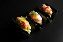 Three pieces of sushi, each topped with different fresh ingredients and garnishes, are arranged on seaweed sheets in a black, wavy sushi holder against a dark background.