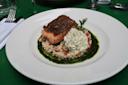 A plated dish featuring seared salmon on a bed of couscous with diced vegetables, topped with a swirl of herbed cream, and surrounded by a green herb sauce on a white plate.