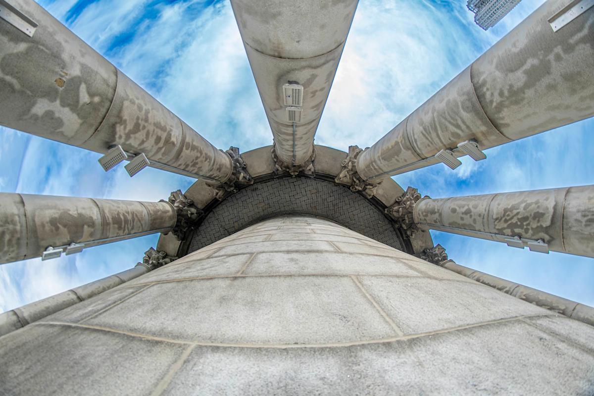 View looking up at several tall, stone columns arranged in a semicircle against a blue sky with clouds, photographed from the base with a wide-angle lens for a dramatic effect.