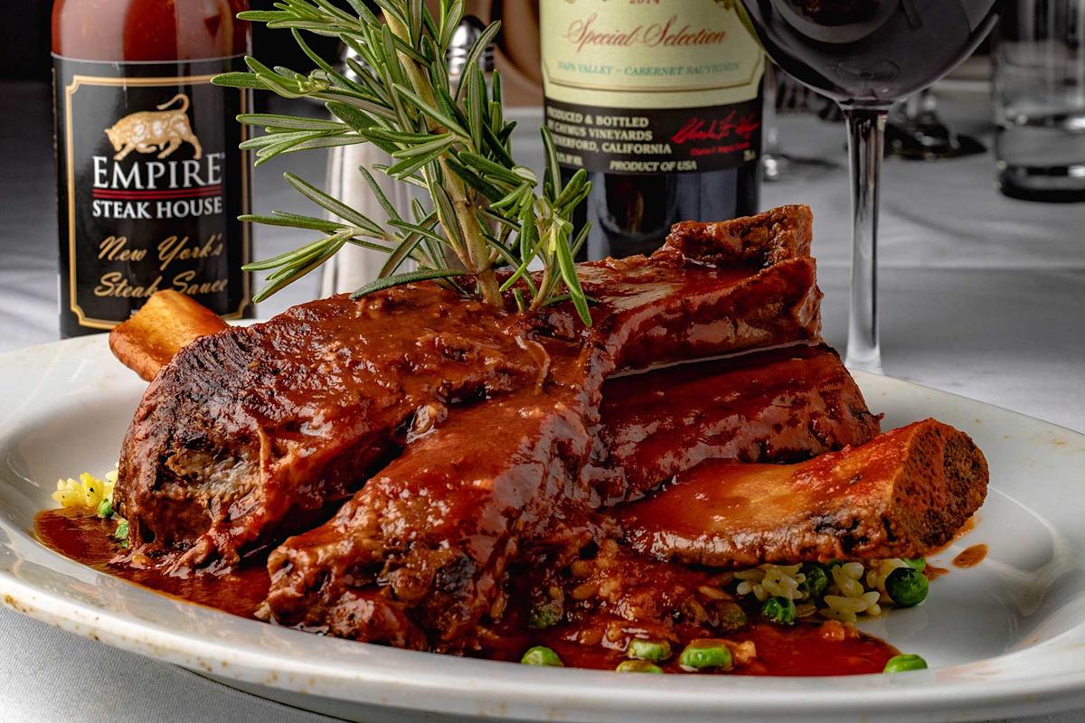 A plate of braised beef ribs in rich brown sauce, garnished with a sprig of rosemary, served over rice and vegetables, with steak sauce and wine bottles and a glass of red wine in the background.