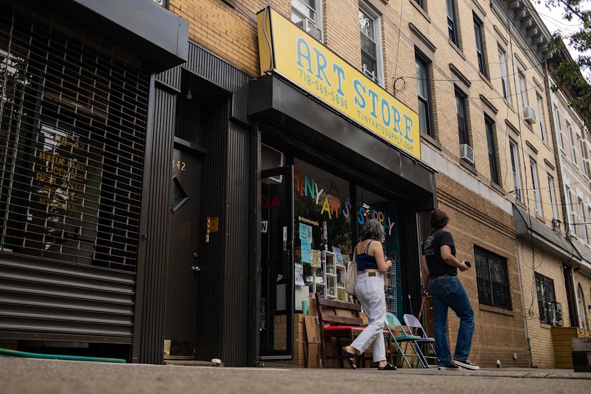 Two people walk past a storefront with a yellow “ART STORE” sign. The shop windows display various art supplies and colorful decorations. The building exterior is tan brick, and the street appears quiet.