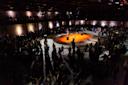 A large crowd gathers in a dimly lit circular venue around an orange-lit central stage with various sculptures. People are standing and walking, with some viewing the art pieces in a spacious, industrial-style setting.