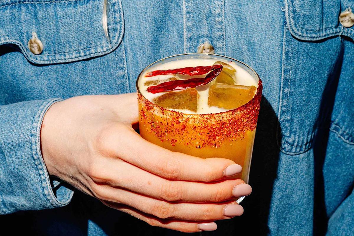 A hand with light pink nails holds a glass of orange cocktail with ice, a chili powder rim, and garnished with dried chili peppers. The person is wearing a blue denim shirt.