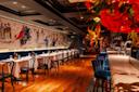 Elegant restaurant interior with wooden floors, tables set with white tablecloths and wine glasses, blue velvet bar stools, a long mural on the wall, and vibrant red flowers in the foreground.