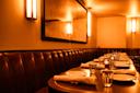 A warmly lit restaurant dining area with a long table set with plates, glasses, and napkins. Brown leather booth seating lines the wall, and modern wall sconces provide ambient lighting. A large mirror hangs above the booth.