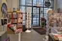 A cozy bookstore with shelves of books, greeting cards on a rotating stand, and assorted gifts. Large windows and a glass door show a city street and scaffolding outside. An exit sign hangs above the door.