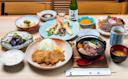 A Japanese meal set on a table, featuring rice, fried cutlets, sautéed vegetables, tofu, soba noodles, tempura, pickles, soup, dipping sauces, green tea, and a bottle of sake with a small cup.