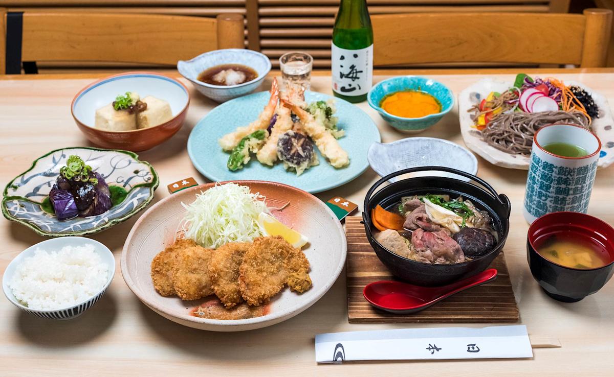 A Japanese meal set on a table, featuring rice, fried cutlets, sautéed vegetables, tofu, soba noodles, tempura, pickles, soup, dipping sauces, green tea, and a bottle of sake with a small cup.