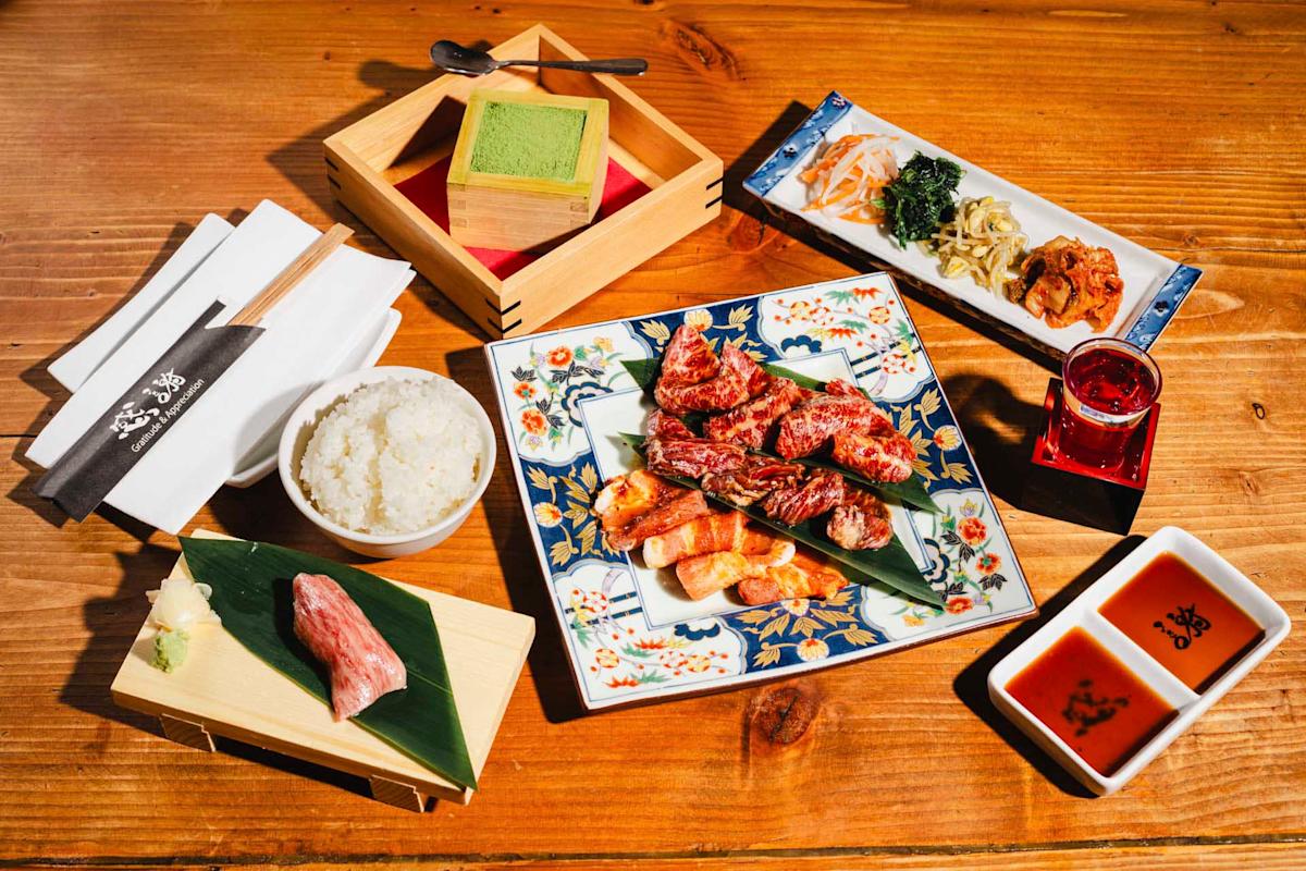 A colorful Japanese meal is arranged on a wooden table. It includes a plate of grilled meat, a bowl of rice, a side dish with greens and kimchi, a piece of meat, a drink, and a wooden box with green matcha powder. Chopsticks rest nearby.