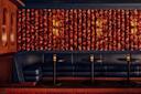 A cozy lounge area features dark blue, curved seating against a patterned red wall. Three round black tables with small candle-lit lamps are arranged along the seating. The warm lighting creates an intimate atmosphere.