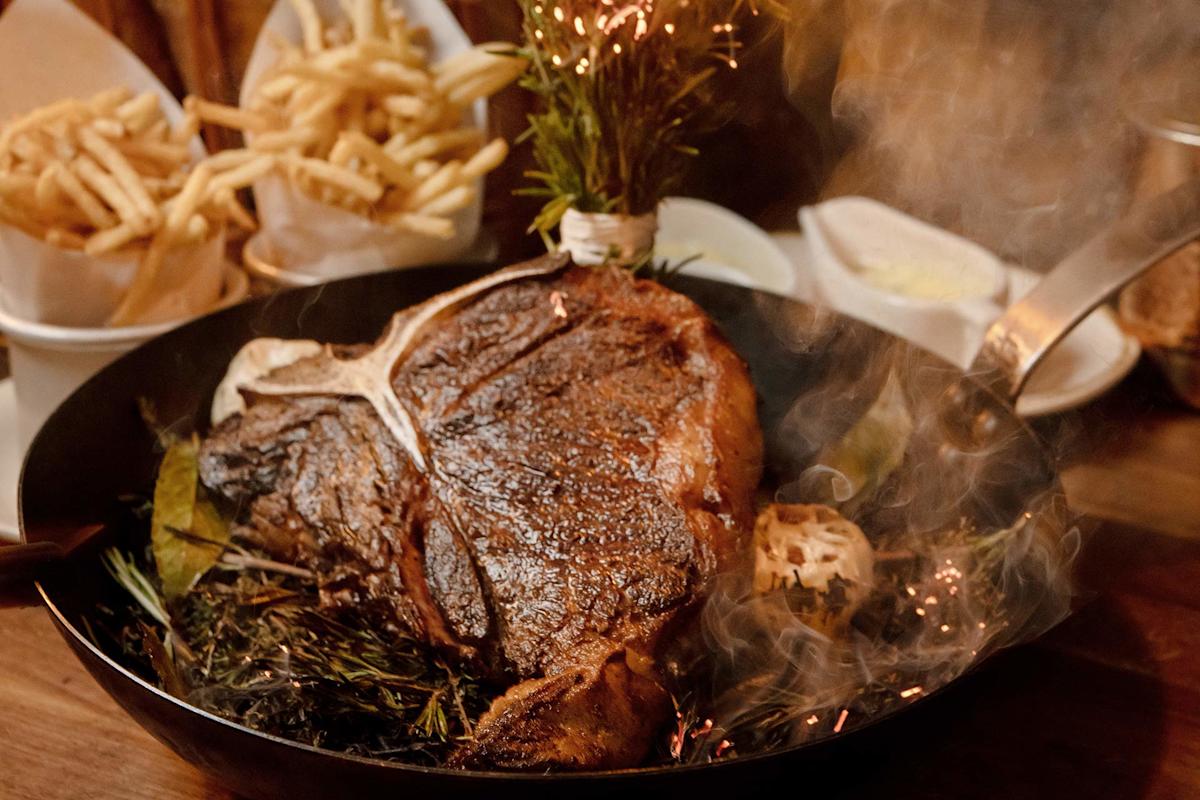 A sizzling, grilled T-bone steak in a skillet with herbs and roasted garlic, with fries in paper cones and a dish of butter in the background. Steam rises from the meat, creating a warm, appetizing scene.