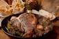 A sizzling, grilled T-bone steak in a skillet with herbs and roasted garlic, with fries in paper cones and a dish of butter in the background. Steam rises from the meat, creating a warm, appetizing scene.
