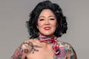 A woman with curly black hair and bright red lipstick smiles confidently. She wears a colorful scarf around her neck and has floral tattoos on her shoulders and upper arms, standing against a plain background.