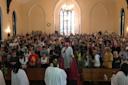 A congregation gathered inside a church with high arched ceilings. A pastor in white robes stands at the front, facing the audience. Stained glass windows illuminate the space. People of various ages are seated and standing, engaged in the service.