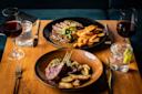 Two elegant plates of food on a wooden table: one with lamb chops and roasted potatoes, the other with steak, fries, and sauce. Two glasses of red wine and two glasses of water with lime are also on the table.