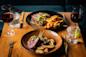 Two elegant plates of food on a wooden table: one with lamb chops and roasted potatoes, the other with steak, fries, and sauce. Two glasses of red wine and two glasses of water with lime are also on the table.
