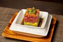 A square stack of diced raw tuna on top of avocado, topped with crispy bits and microgreens, sits in soy sauce on a white dish over ice, presented on a wooden tray.