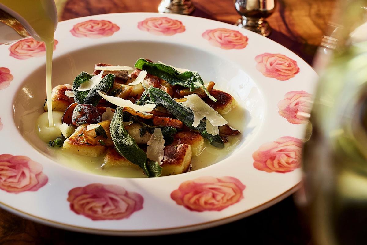 A plate with flower patterns holds gnocchi topped with sage, mushrooms, and shaved cheese, while a creamy sauce is being poured over the dish.