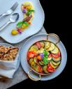 A dish of ratatouille with sliced tomatoes, zucchini, and eggplant, garnished with basil and edible flowers, is served alongside grilled bread and an elegant plated appetizer with colorful garnishes.