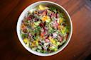 A colorful salad in a white bowl, topped with microgreens, capers, radishes, tomatoes, cucumbers, yellow peppers, and red onions, set on a wooden table.
