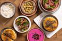 Assorted Middle Eastern dishes including pita, hummus, labneh, beet dip, tabbouleh, and baba ganoush in colorful bowls on a wooden table, with part of a menu visible.