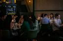 A group of people sit at tables in a dimly lit restaurant, engaged in conversation and eating. The scene is cozy, with warm lighting and a relaxed atmosphere.