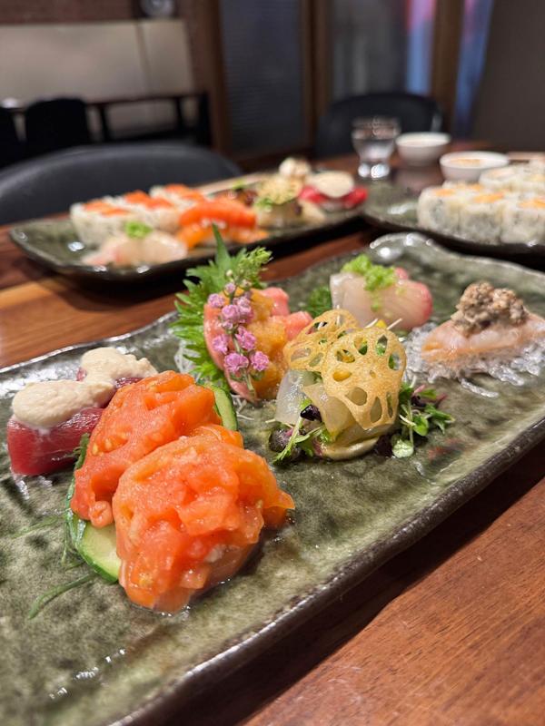 A close-up of assorted sushi and sashimi artfully arranged on a rectangular ceramic plate, garnished with edible flowers, greens, and lotus chips, with more sushi plates blurred in the background on a wooden table.