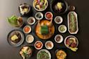 A diverse spread of Korean dishes on a dark table, featuring a whole chicken, various small side dishes, soups, noodles, and grilled items. Some dishes include fresh greens, vegetables, and garnishes.