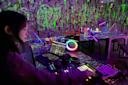 A person operates DJ equipment and a laptop in a dimly lit room with neon art supplies and fluorescent paint splatters on the walls and tables, creating a vibrant, creative atmosphere.