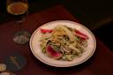 A plated Caesar salad with slices of watermelon radish and shredded cheese on a dark wooden table. There is a glass of white wine and a coaster nearby.