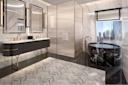 Luxurious bathroom with marble walls and floors, a double sink vanity, large mirrors, a round black marble bathtub, gold fixtures, and a glass shower enclosure. A window offers a city skyline view.