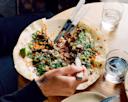 A person holds a fork over a loaded flatbread topped with greens, meat, and sauce on a wooden table with two glasses of water and utensils.