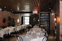 A dimly lit restaurant with neatly set tables covered in white linens. Black chairs surround the tables, and the décor features exposed brick walls, a patterned floor, and a tin ceiling. A small bar area is visible in the background.