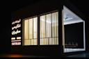 A modern, illuminated building with large windows and beige curtains. Neon Persian text and "SAFIRAN" are displayed on the wall. Several empty chairs are visible inside under bright lights. The setting is dark outside.