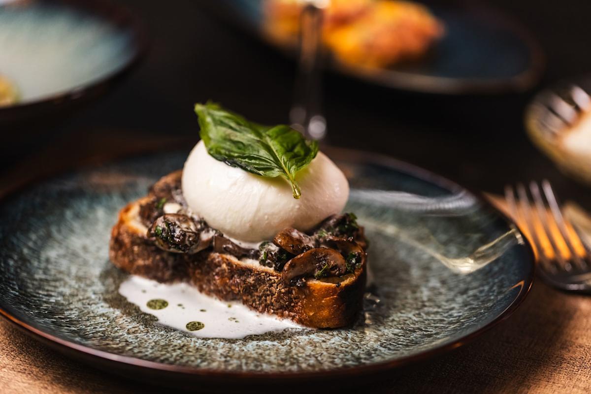 A slice of toasted bread topped with mushrooms, a poached egg, creamy sauce, and a fresh basil leaf is served on a blue plate with a fork beside it.