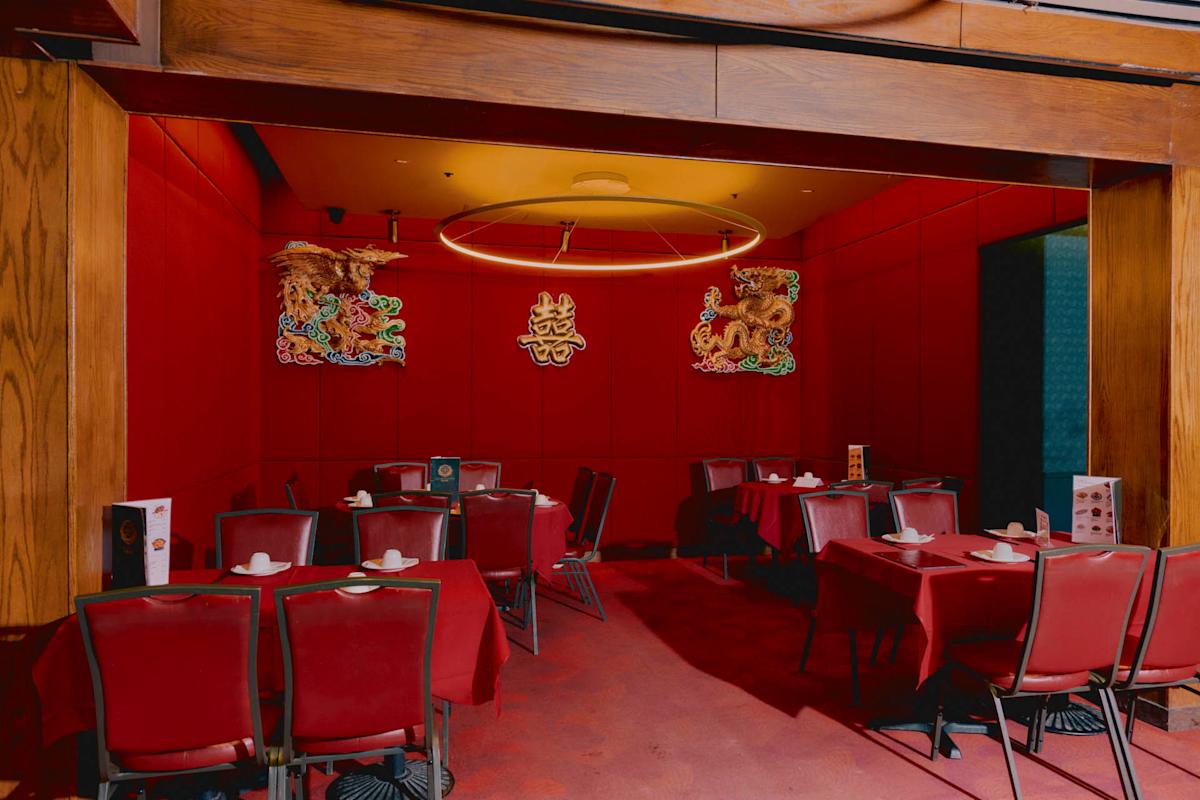 A dimly lit restaurant with red walls and tablecloths, set with plates and chopsticks. Decorative Chinese dragon and phoenix wall art, along with a circular light fixture, hang above the tables.