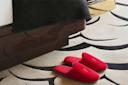 A pair of red slippers rest on a patterned beige and black rug next to a wooden bed frame with a black blanket draped over the edge.