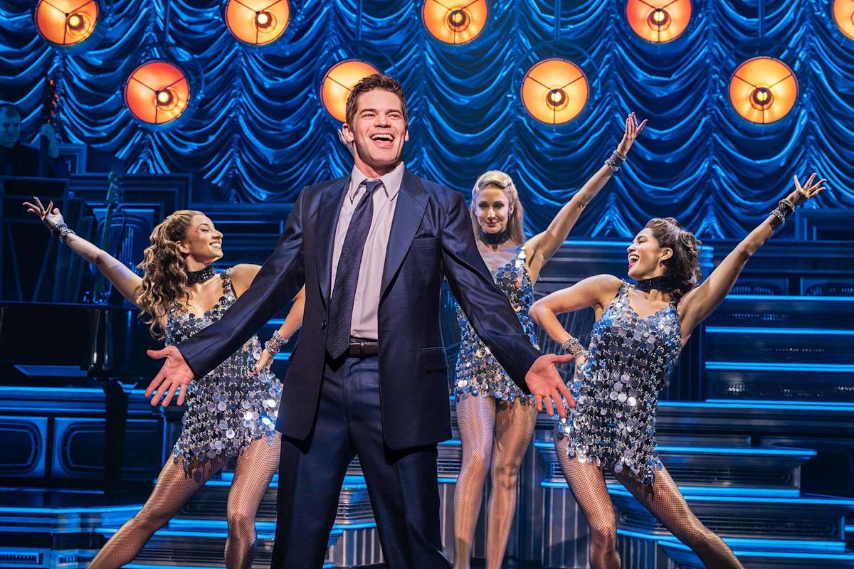 A man in a suit stands center stage singing with a big smile, arms wide open. Three women in sparkly dresses dance behind him. Bright lights and blue drapery create a lively, theatrical atmosphere.