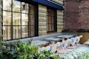 Outdoor cafe patio with patterned tables, white chairs, and a long gray cushioned bench against a textured wall with geometric tiles. Brick wall and greenery add to the cozy, modern atmosphere.