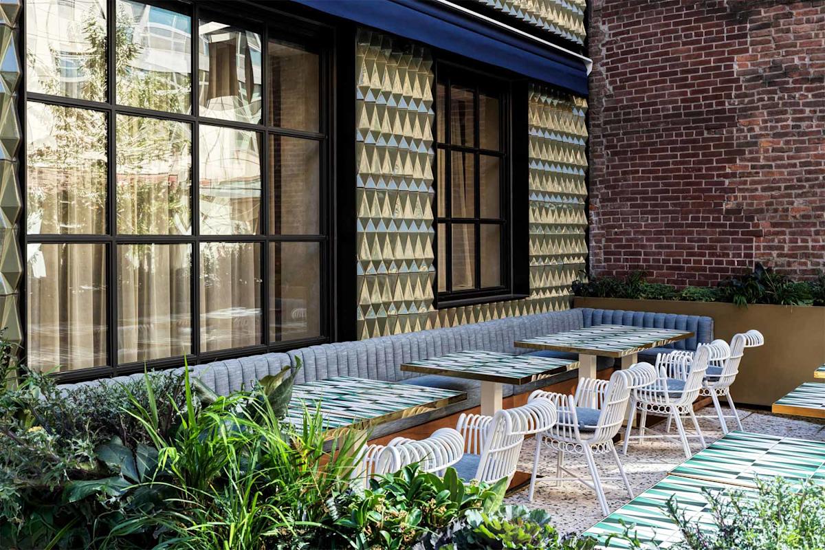 Outdoor cafe patio with patterned tables, white chairs, and a long gray cushioned bench against a textured wall with geometric tiles. Brick wall and greenery add to the cozy, modern atmosphere.