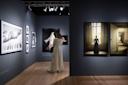 A person in a white coat admires art in a dimly lit gallery. The walls display various large framed photographs, including monochrome and color images. Track lighting highlights the artwork. The floor is wooden.
