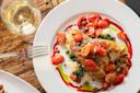 A plate of grilled fish topped with sautéed cherry tomatoes, served over sautéed greens and creamy grits, garnished with capers and colorful sauce, next to a glass of white wine on a wooden table.