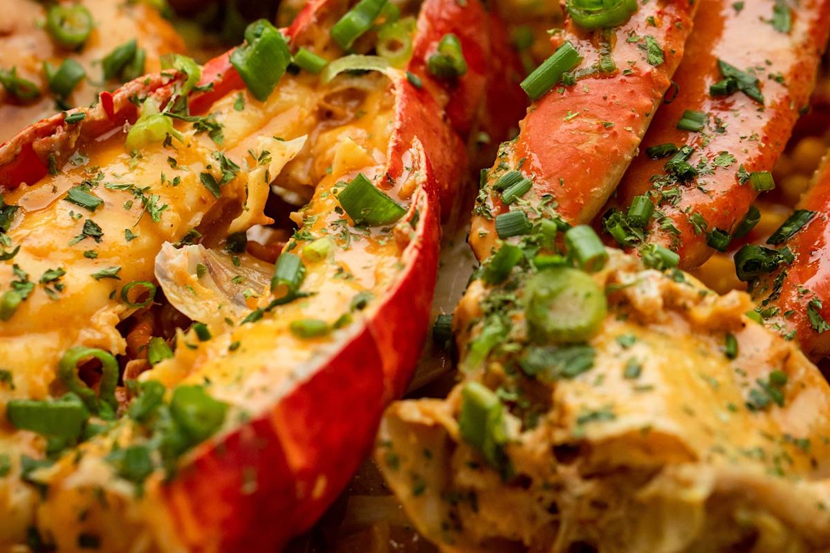 Close-up of a cooked lobster dish topped with melted cheese, chopped green onions, and herbs, highlighting the vibrant reds of the lobster shell and the fresh green garnishes.